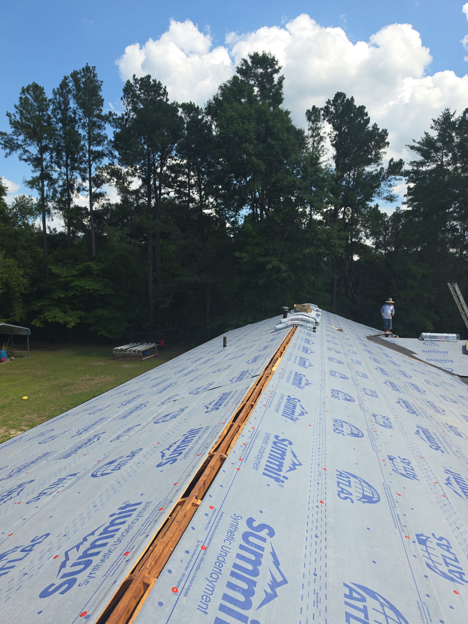 Synthetic underlayment rolled tight across the repaired deck and the ridge slot cut clean, with shingle bundles staged and ready to go on