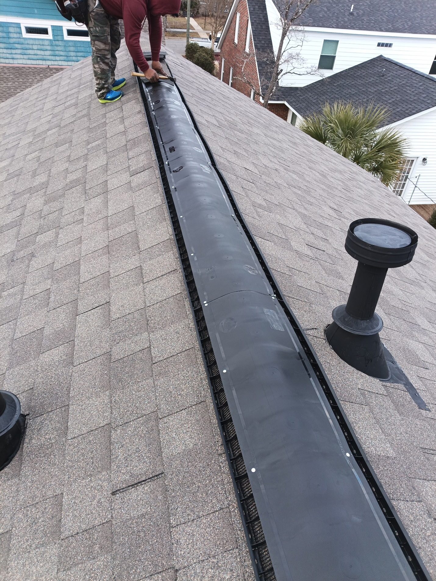 A crew member bedding the ridge vent into the new shingle field, with a fresh pipe boot set on the